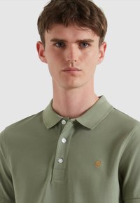 Hellgrünes Polohemd aus Baumwolle. Verfügt über eine Knopfleiste mit drei Knöpfen und ein kleines gesticktes Logo in Orange auf der Brust.