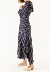 Kobieta w granatowo-beżowej w kratę sukience z długim rękawem o asymetrycznym zupełnieniu, zestawionej z beżowymi butami na obcasie w kształcie slip-on.