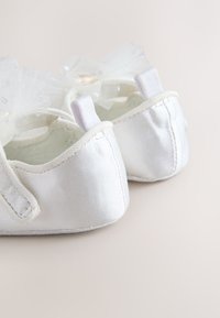 Scarpette da balletto in raso bianco con fiocchi in tulle trasparente, cinturini elastici sul tallone e finitura liscia. Presentano cuciture sottili lungo i bordi.