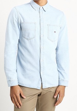 Man draagt lichtblauw denim overhemd met knopen en twee borstzakken, en beige broek, staat met ontspannen handen langs zijn zij.