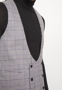 Gekachelte Weste in Grau und Marineblau, mit tiefem V-Ausschnitt, sechs Knöpfen und einer einzelnen Brusttasche; kombiniert mit einem schwarzen Langarmoberteil.