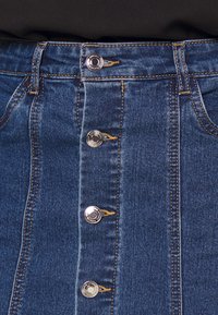 Närbild av en blå denimkjol med vertikala silverknappar och orange sömmar på en svart bakgrund.