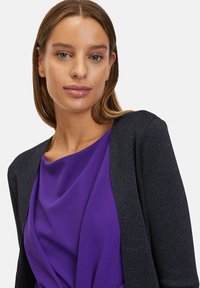 Fekete, csillogó kardigán egy lila, laza blúz fölött. Sima szövettextúrák, karcsú fazon, élénk színkontrasztot mutatva.