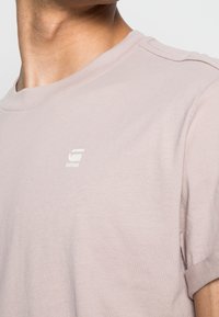 Detailní záběr na krk a rameno oblečené v světle růžovém tričku s malým bílým logem "RAW" na hrudi.