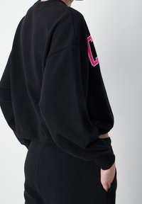 Svart cropped sweatshirt med en rosa grafisk bokstav på framsidan, långa ärmar och ribbade mudd, tillverkad av mjukt bomullsblandat material.