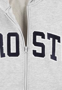 Szara bluza z zamkiem, z wyraźnymi granatowymi literami "PRO ST" z przodu, prezentująca wyszywaną teksturę i detal zamka.