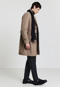 Soprabito di lana beige con grandi bottoni e tasche laterali, abbinato a una sciarpa nera e pantaloni grigi, completato da scarpe nere con lacci.