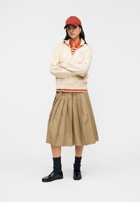 Μπεζ φούτερ με φερμουάρ, πορτοκαλί και λευκό ριγέ πόλο, χακί πλισέ φούστα, μαύρα παπούτσια, ναυτικό μπλε υφασμάτινες κάλτσες, κόκκινο καπέλο.