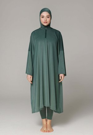 Vrouw staat blootsvoets en draagt een volledig bedekkende bescheiden badkledingset in donkergroen met lange mouwen en hijab.