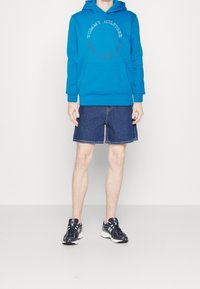 Sudadera con capucha azul con un bolsillo frontal y texto con logotipo, combinada con pantalones cortos de mezclilla y zapatillas azul marino con detalles en blanco.