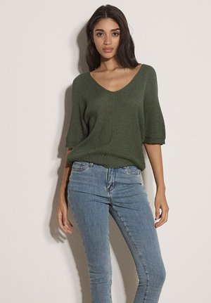 Kobieta ubrana w luźny oliwkowozielony sweter z dzianiny z rękawami do łokcia i jasnoniebieskie dopasowane dżinsy, stojąca na tle jasnej, gładkiej ściany.