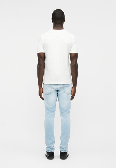 Man standing facing away, wearing a plain white short-sleeve t-shirt, light blue jeans, and black sneakers, against a white background.