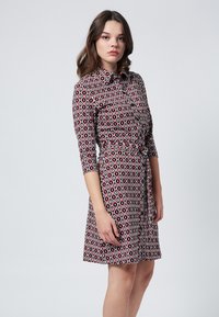 Paita kaulalla, jossa on pitkät hihat, ja geometrinen punainen, musta ja valkoinen kuosi. Siinä on vyötärönauha ja liehuva hame. Sileä kangas.