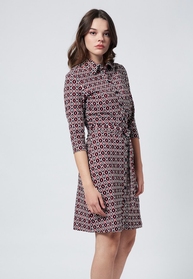 Paita kaulalla, jossa on pitkät hihat, ja geometrinen punainen, musta ja valkoinen kuosi. Siinä on vyötärönauha ja liehuva hame. Sileä kangas.