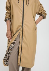 Manteau léger beige avec fermetures éclair noires, doté d'une doublure et d'accents de poignet en imprimé léopard. Design long avec des poches minimalistes.