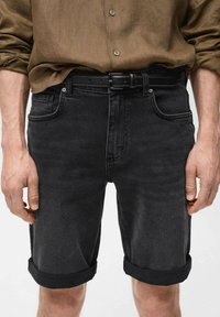 Schwarze Denim-Shorts mit umgeschlagenen Säumen, ausgestattet mit einem Knopfverschluss, fünf Taschen und gestickten Details, kombiniert mit einem braunen knöpfbaren Hemd.