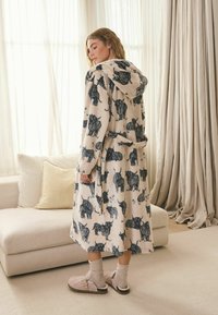 Nő áll bent, bézs színű kapucnis köntöst visel fekete tehénmintákkal, gyapjúzoknit és rózsaszín papucsot egy lágyan megvilágított nappaliban.