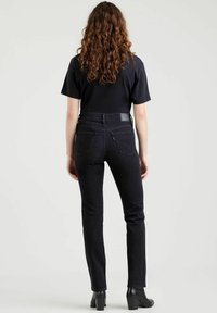 Jeans denim hitam dengan desain kaki lurus, dilengkapi saku belakang dan label di pinggang. Dipadukan dengan atasan hitam lengan pendek.