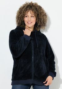 Kvinde med krøllet hår iført en marineblå fuzzy zip-up hoodie og hoops-øreringe, smilende og kiggende til siden mod en hvid baggrund.
