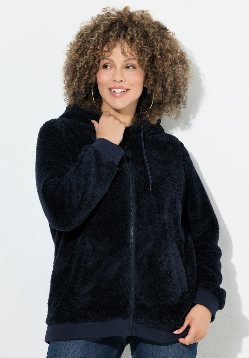 Kvinde med krøllet hår iført en marineblå fuzzy zip-up hoodie og hoops-øreringe, smilende og kiggende til siden mod en hvid baggrund.