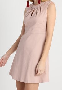 Abito senza maniche di un rosa chiaro testurizzato con un dettaglio sul collo a chiave indossato da una persona con capelli lunghi e orecchini a frangia rossi.