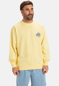 Yellow sweatshirt with a round neckline and long sleeves. Features a blue and gray triangular logo on the left chest. Soft fabric texture.
