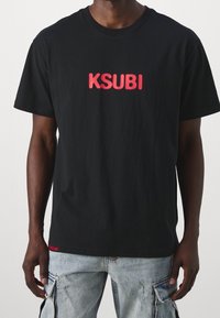 Černé bavlněné tričko s odvážným červeným vyšitým logem "KSUBI" na přední straně a malou červenou nášivkou s nápisem "SAYS ME" na lemu.
