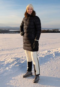 Svart isolerad pufferjacka, krämfärgade byxor med svarta sidor och grå stövlar. Personen har en beige stickad mössa och svarta handskar.