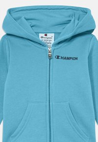 Modrá zipsová mikina z mäkkého materiálu s kapucňou. Obsahuje predné vrecká a čierne logo "CHAMPION" na hrudi. Veľkosť: 12 mesiacov.