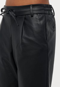 Zwarte leren broek met een gladde textuur, geplooid ontwerp, elastische tailleband en verstelbaar trekkoord. Heeft zijzakken.