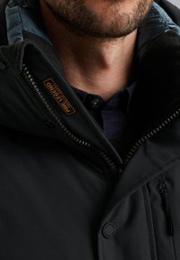 Zwarte jas met een hoge kraag, voorzien van een rits aan de voorkant, textuurstof en een badge met het label "PINE LEGEND." De binnenkant is gevoerd met blauw.