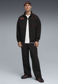 Sort atletisk træningsdragt med røde accenter og et logo, bestående af en lynlåsjakke og løstsiddende bukser, parret med sportssko.