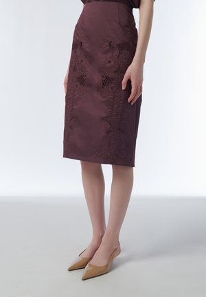 Woman wearing a knee-length dark purple skirt with floral embroidery and beige pointed-toe low heels, standing on a light floor.