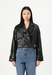 Veste en simili cuir noir coupée avec un motif texturé, larges revers, grande poche avant et taille ceinturée, associée à un jean bleu clair.