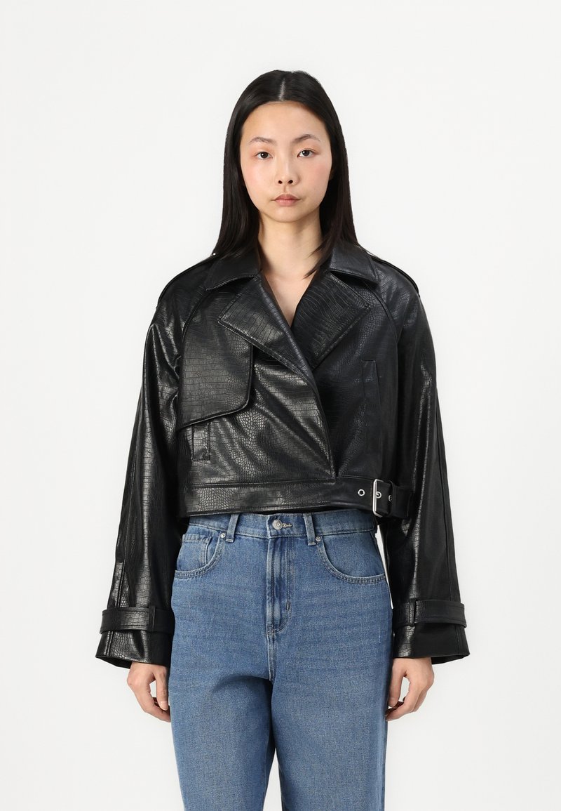 Veste en simili cuir noir coupée avec un motif texturé, larges revers, grande poche avant et taille ceinturée, associée à un jean bleu clair.