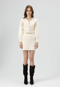 CrÈmekleurige truienset met een V-hals en drie grote knopen, gecombineerd met een bijpassend mini-rokje. Model draagt donkerbruine hoge laarzen tot de knie.