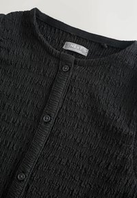 Camicia nera a bottoni con trama, scollo rotondo e etichetta "NEXT Made in Sri Lanka" all'interno del colletto.