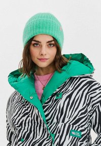 Prošívaná bunda s černo-bílým zebrovým vzorem, zelená podšívka kapuce, doplněná o žebrovanou mint zelenou čepici. Pod ní světle růžový svetr.