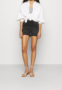 Black denim shorts with frayed hem, paired with a knotted white button-up shirt. Footwear includes white strappy sandals with flat soles.