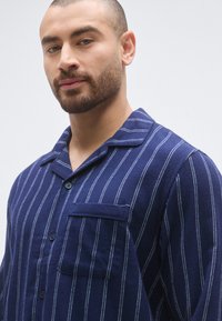 Granatowa koszula flanelowa w paski, z kołnierzykiem na guziki, przednią kieszenią i dwoma guzikami, z białymi pionowymi paskami dla kontrastu.