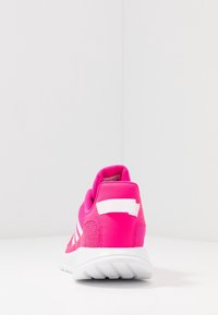 Zapatilla deportiva de color rosa brillante con franjas blancas y suela blanca, vista desde atrás sobre una superficie y fondo blancos.