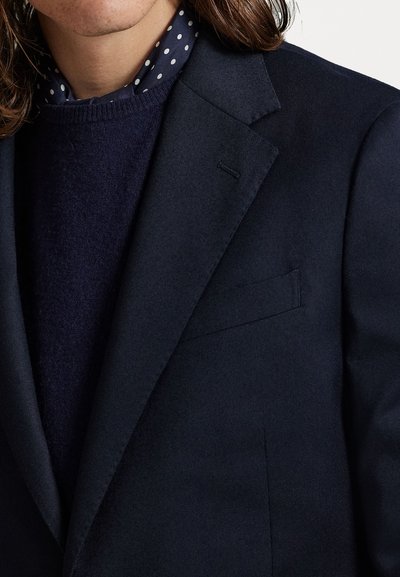 Blazer azul-marinho com uma textura suave, lapela simples, e um bolso no peito, sobre um suéter de gola redonda azul-marinho escuro e uma camisa com padrões de bolinhas.
