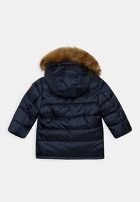 Chaqueta acolchada azul marino con capucha desmontable, que presenta un borde de piel sintética marrón y una textura acolchada. Mangas cortas y sin cierre frontal visible.