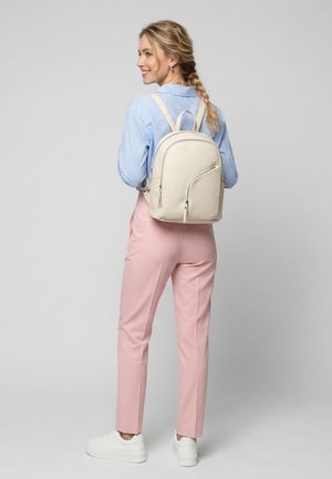 Kvinde med blond flet iført blåternet skjorte, lyserøde bukser, hvide sneakers og med en cremefarvet rygsæk, smilende og kigger til siden.