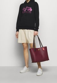Sudadera negra con gráfica rosa, pantalones cortos plisados beige y un bolso de cuero burdeos con asas marrones y una etiqueta. Zapatillas blancas.