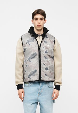 Noor mees, kes kannab beeži pusa, helesiniseid teksasid ja halli mustrilist lukuga vestiga, millel on must krae, seisab ühevärvilise tausta ees.