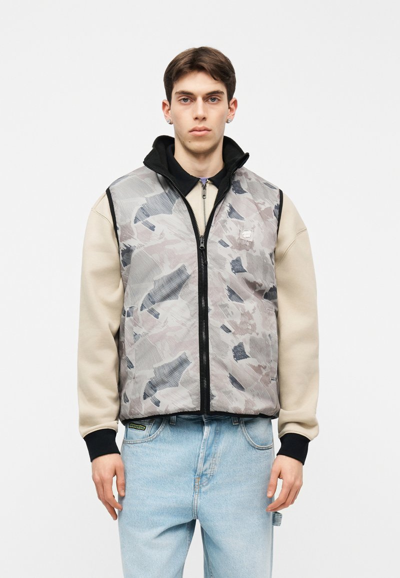 Νέος άνδρας με μπεζ φούτερ, ανοιχτόχρωμα μπλε τζιν και γκρι γιλέκο με φερμουάρ και μαύρο γιακά, στέκεται μπροστά σε απλό φόντο.