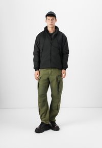 Veste zippée noire isolée, pantalons cargo verts avec poches, bottes noires épaisses et une casquette sombre, se tenant devant un fond uni.