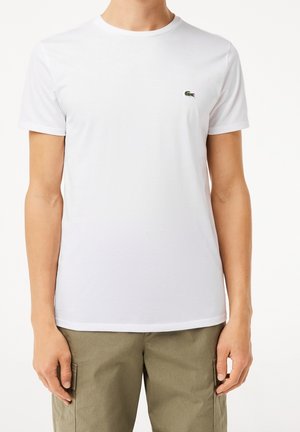 Mann trägt ein schlichtes weißes T-Shirt mit kurzen Ärmeln und einem kleinen grünen Krokodil-Logo auf der Brust sowie olivgrüne Cargohosen.