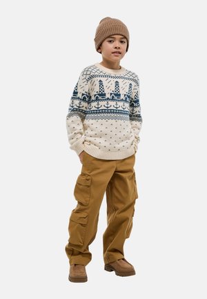 Garçon portant un bonnet beige, un pull crème avec un motif de phare bleu marine, un pantalon cargo marron et des bottes beige clair, debout les mains dans les poches.